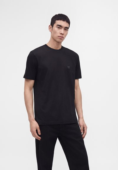 Jeune homme aux cheveux noirs courts portant un t-shirt noir uni à manches courtes et un pantalon noir sur fond blanc.