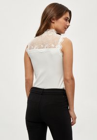 Top bianco senza maniche con dettagli in pizzo sulla schiena; tessuto liscio; orli sagomati. Abbinato a pantaloni neri attillati.