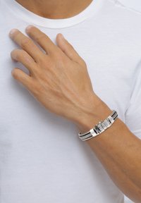 Armband aus silberfarbenem Metall mit strukturiertem Glieddesign und einem Verschluss, der an einer Hand vor einem weißen T-Shirt-Hintergrund getragen wird.