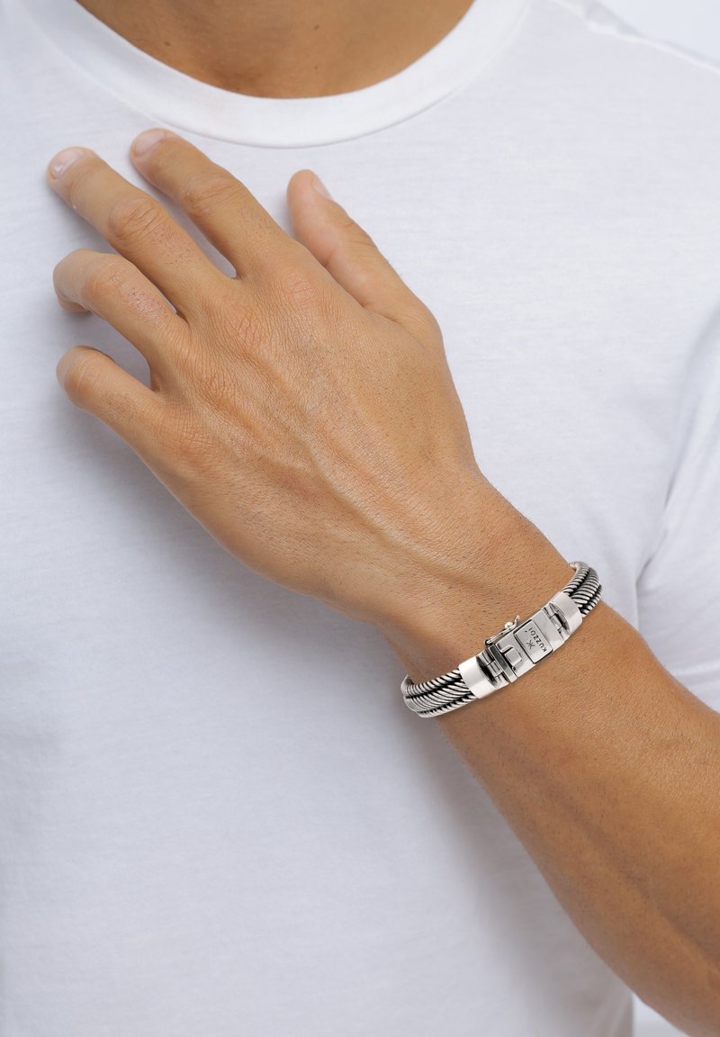 Armband aus silberfarbenem Metall mit strukturiertem Glieddesign und einem Verschluss, der an einer Hand vor einem weißen T-Shirt-Hintergrund getragen wird.