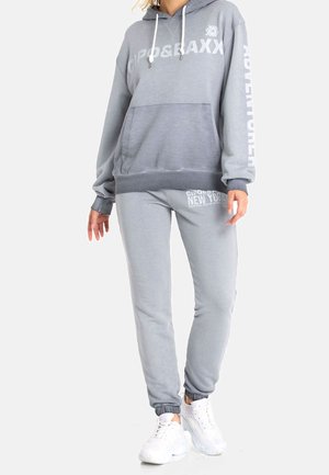 Person iført en lysegrå hættetrøje med frontlomme og matchende joggingbukser, begge med hvidt trykt tekst, kombineret med hvide sneakers.