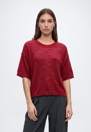 Vrouw met steil bruin haar draagt een getextureerde rode korte mouwen top en donkere geplooide broek, staand tegen een effen lichte achtergrond.
