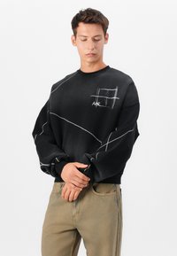 Schwarzer Sweatshirt mit weißen Nahtdetails und abstraktem Grafikdesign auf der Brust. Lockerer Schnitt und gerippte Bündchen. Trägt man mit khakifarbenen Hosen.