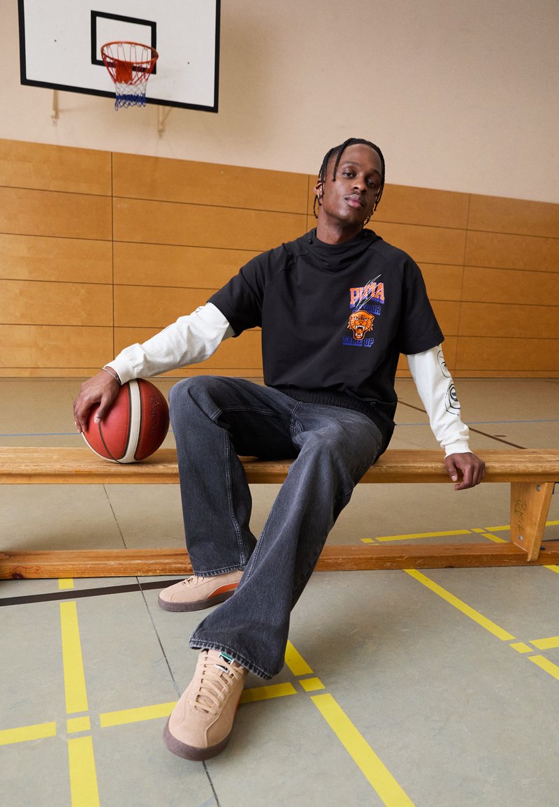 Sudadera negra con gráfico naranja, superpuesta sobre una camiseta blanca de manga larga. Pantalones de mezclilla oscuros y zapatillas beige, sentado en un banco con un balón de baloncesto.