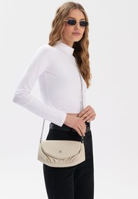 Beige læderhåndtaske med kædestrop, der har et buet klapdesign og tekstureret overflade, holdt af en person iført en hvid top og sorte bukser.