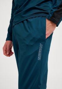 Aqua sportieve broek gemaakt van glad materiaal, met een elastische tailleband en zijzak, met het 'HUMMEL' logo verticaal in het wit gedrukt.