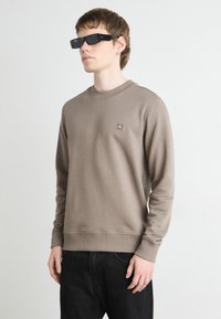 Sudadera marrón con cuello redondo y puños de canalé, que presenta un pequeño parche con logotipo en el pecho. Combinada con gafas de sol negras elegantes y jeans oscuros.