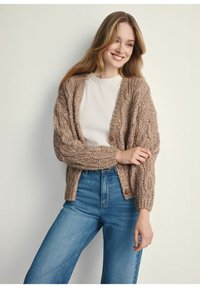 Brązowy sweter z dzianiny z wzorem w warkocze, o luźnym kroju, z dekoltem w serek i drewnianymi guzikami, w połączeniu z jasnoniebieskimi spodniami z wysokim stanem.