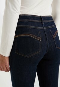 Persona que lleva jeans de mezclilla azul oscuro con costuras en contraste y una camiseta blanca de manga larga, visto desde la cintura hacia abajo por la parte de atrás.