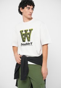T-shirt branco com o logótipo texturizado verde "W" e gráfico "Double A". O modelo usa um suéter escuro amarrado na cintura, combinado com calções de carga verdes.