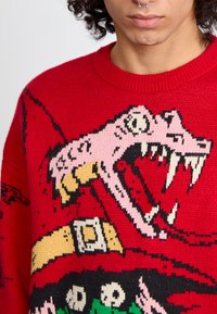 Suéter de punto rojo con un diseño gráfico de una gran criatura de boca abierta en rosa y negro, con detalles como un cinturón amarillo.