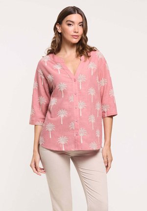 Mujer con blusa rosa abotonada con estampados blancos de palmeras y pantalones beige, de pie contra un fondo claro y liso.