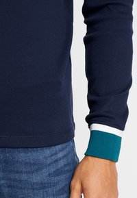 Prawa ręka i tors w granatowej koszuli z długim rękawem z zielonym i białym mankietem, w połączeniu z niebieskimi dżinsami, na jednolitym tle.