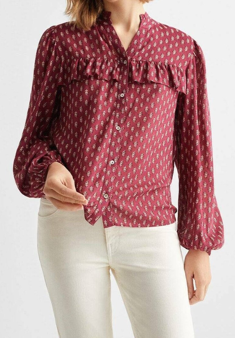 Femme portant une blouse rouge à motifs avec des volants et un pantalon blanc, en train de boutonner la blouse sur le devant.