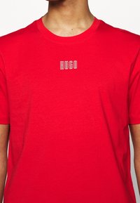 Person iført en rød T-shirt med rund hals og hvidt omrids tekst "HUGO" centreret på brystet.