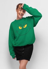 Felpa verde con stampa del volto del Grinch e occhi gialli, abbinata a una gonna di jeans nera con dettagli a bottoni e una silhouette aderente.