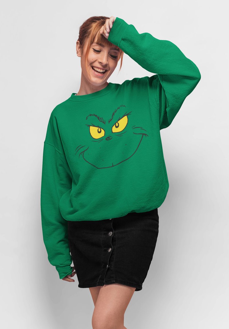 Felpa verde con stampa del volto del Grinch e occhi gialli, abbinata a una gonna di jeans nera con dettagli a bottoni e una silhouette aderente.