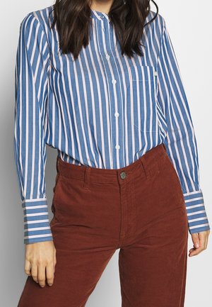 Woman wearing a blue and white vertical striped button-up shirt tucked into high-waisted rust-colored corduroy pants.