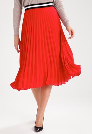 Vrouw draagt een felrode geplooide midirok met een zwart, wit en rood tailleband, gecombineerd met zwarte hoge hakken.