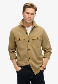 Chemise marron à boutons avec deux poches poitrine, boutons noirs, manches retroussées, et un sous-vêtement blanc, associée à un jean bleu foncé.