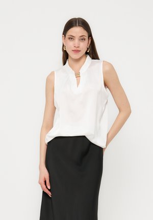 Kvinne med langt brunt hår, iført ermeløs hvit bluse og svart skjørt, accessorert med gullfargede hoop-øredobber og halskjede, står foran hvit bakgrunn.