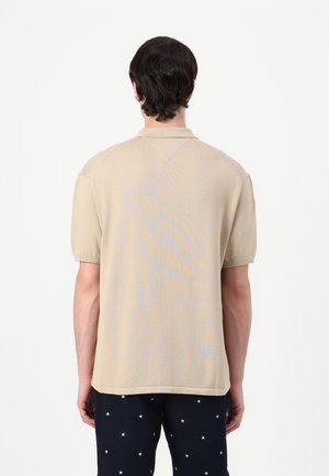 Homme portant un pull en maille beige à manches courtes et un pantalon foncé avec des motifs d'étoiles blanches, vu de dos.