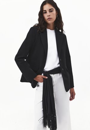 Woman wearing a black blazer over a white shirt with white pants, accessorized with a black fringed scarf tied at the waist.