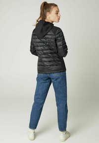 Zwarte pufferjack met een slim fit ontwerp, gewatteerde textuur en een capuchon, gecombineerd met blauwe jeans en witte sneakers, gezien vanaf de achterkant.