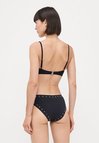 Bikini noir avec des fines bretelles, présentant une fermeture à crochet à l'arrière et des clous métalliques décoratifs autour de la taille haute.