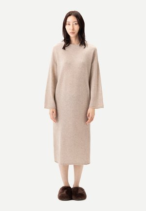 Femme portant une robe beige en maille à manches longues et des chaussons bruns duveteux, debout devant un fond blanc uni.
