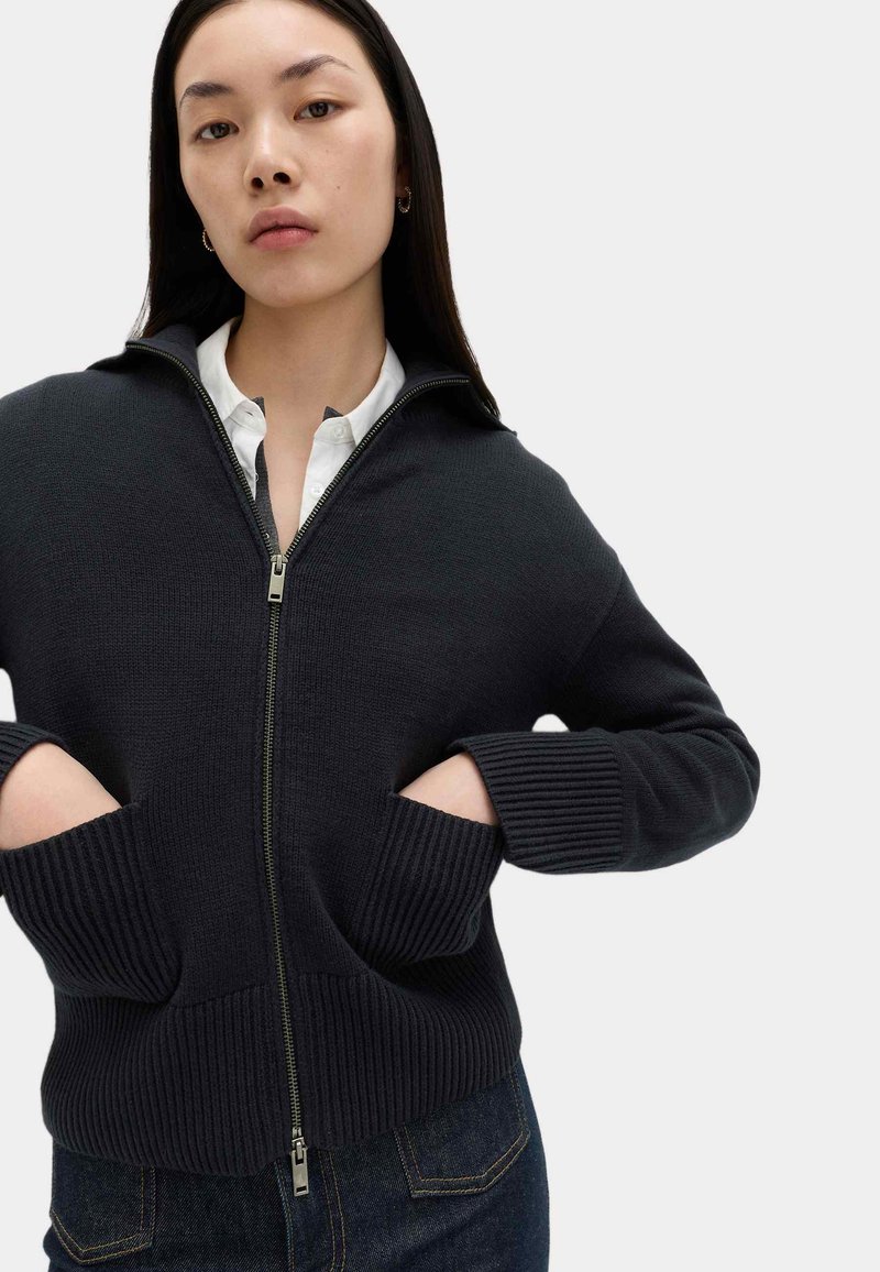 Jeune femme aux cheveux noirs raides portant un pull à fermeture éclair foncé, les mains dans les poches avant, sur une chemise blanche.