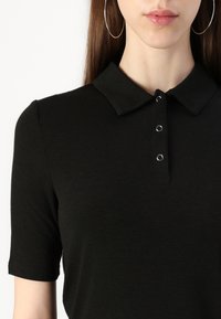 Melna polo krekls ar apkakles izgriezumu un īsām piedurknēm. Funkcijas: trīs metāla pogcaurumi apkaklē. Mīksta, gluda tekstūra.