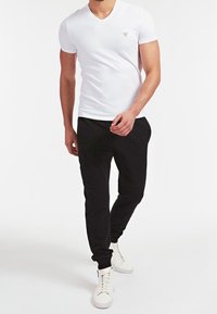 T-shirt bianco a scollo a V con un piccolo emblem, abbinato a pantaloni jogger neri e sneakers bianche. Design semplice e aderente, con una texture liscia.