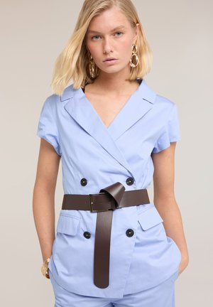 Blonde Frau trägt hellblaues kurzärmeliges Blazer-Kleid mit schwarzen Knöpfen, breitem dunkelbraunem Gürtel und goldenen Creolen.