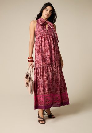 Donna che indossa un abito lungo rosa senza maniche con fantasia floreale e collo halter, tiene una borsa beige, accessoriata con braccialetti rossi e tacchi neri.