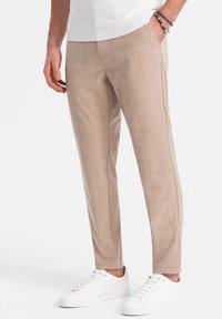 Un pantalon à carreaux beige en tissu léger, coupe slim, avec un motif à quadrillage subtil, associé à des baskets blanches. Design cropped au niveau des chevilles.