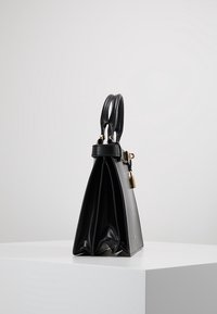 Borsa a mano in pelle nera con doppi manici e dettaglio in oro, in piedi su una superficie bianca contro uno sfondo semplice.