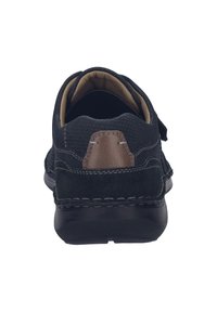Scarpa nera in suede e materiale sintetico con un dettaglio in pelle marrone sul tallone, cuciture visibili e una parte superiore testurizzata. Suola piatta in gomma.