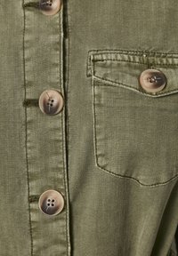 Olivově zelená košile s texturovanou tkaninou, se čtyřmi kulatými světle zbarvenými knoflíky a malou kapsičkou na levé části hrudi.