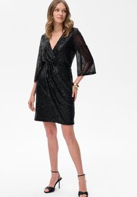 Kobieta w czarnej sukience z cekinami, z przezroczystymi rękawami i czarnymi sandałami na wysokim obcasie, pozująca na białym tle.