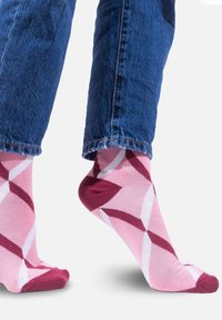 Różowo-bordowe skarpety w wzory z białymi kształtami geometrycznymi, wykonane z miękkiego materiału, noszone z niebieskimi dżinsami.