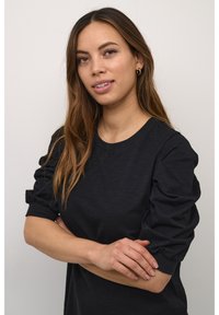 Melna T krekls ar apaļu kakla izgriezumu un uzpūstām piedurknēm, ar mīkstu tekstūru. Modelis sakrustojusi rokas uz neitrāla fona.