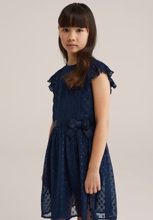 Niña joven con cabello largo, vestida con un vestido azul marino con mangas con volantes y un cinturón de lazo, posando contra un fondo liso.