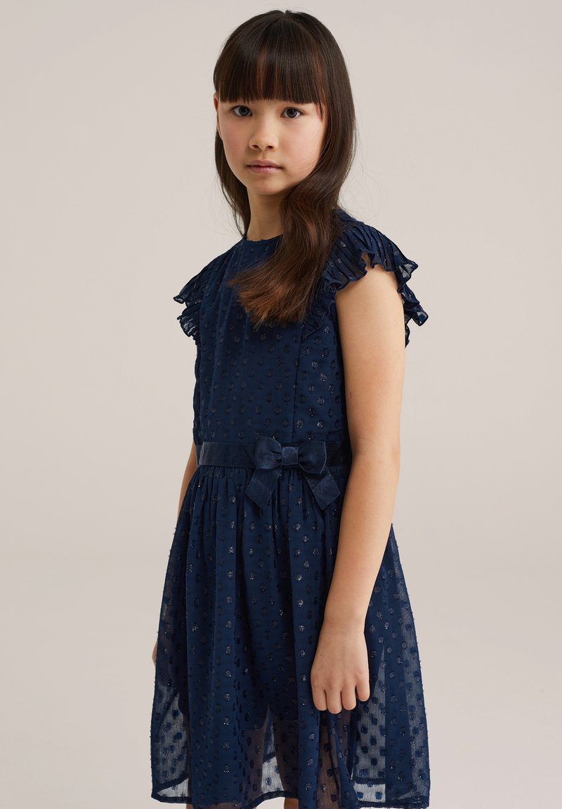 Hosszú hajú fiatal lány, sötétkék, fodros ujjú ruhában, masnis övvel, egyszínű háttér előtt áll.