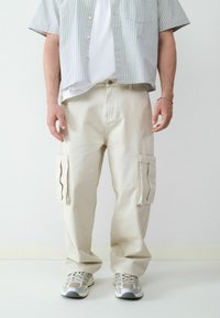 Beige cargobroeken met zijzakken, gemaakt van stevige stof, gecombineerd met een wit T-shirt en een kortlopende geruite shirt. Lichtgekleurde sneakers.