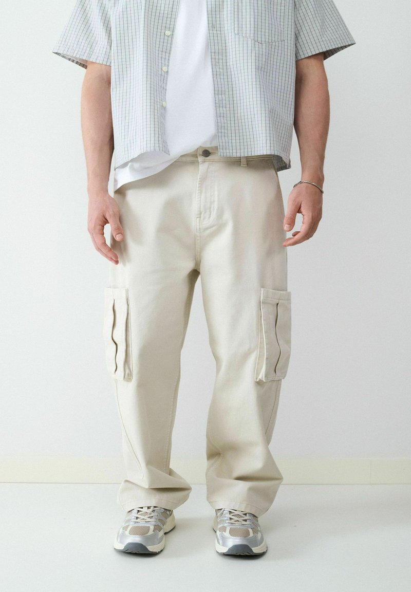 Beige cargobroeken met zijzakken, gemaakt van stevige stof, gecombineerd met een wit T-shirt en een kortlopende geruite shirt. Lichtgekleurde sneakers.