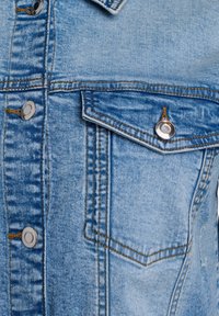 Šviesiai mėlynas džinsinis švarkas su sidabriniais spaudimo mygtukais, nusidėvėjusia tekstūra ir krūtinės kišene. Pasižymi kontrastinga siuvimo linija ir smailiu apykakle.