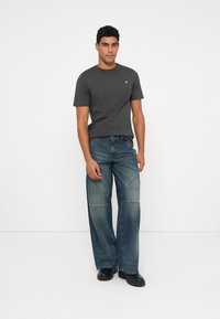 T-shirt de algodão cinza com logótipo amarelo, combinado com jeans de brim azuis de corte solto, apresentando um design de remendos e costura em contraste. Sapatos pretos.