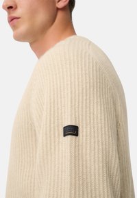 Beige gestreifter Strickpullover mit einem strukturierten Muster, versehen mit einem schwarzen Etikett auf der linken Schulter. Das Design hat einen Rundhalsausschnitt und lange Ärmel.