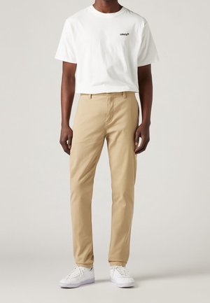 T-shirt Levi's à manches courtes blanche avec logo noir, associé à un pantalon beige clair et des baskets blanches, créant un look décontracté.